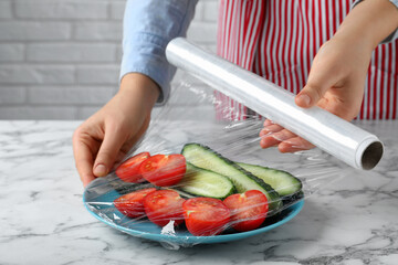 Wall Mural - Woman putting plastic food wrap over plate of fresh vegetables at white marble table indoors, closeup