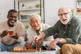 Fototapeta  - Cheerful multiethnic elderly friends looking at camera near chess and tea at home.