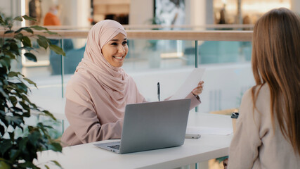 Wall Mural - Young arab woman consultant sales agent bank worker sitting at office desk advises unrecognizable girl client offers to sign contract sale agreement helps to arrange loan proposes legal consultation