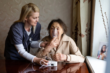 Wall Mural - Young high-skilled pleasant woman doctor putting on nebulizer mask on face of her elderly female patient to make inhalation when visitng him at home. Flu, cold and cough treatment.