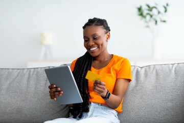 Joyful black woman making online payment, buying goods on web, using tablet and credit card at home. Internet shopping