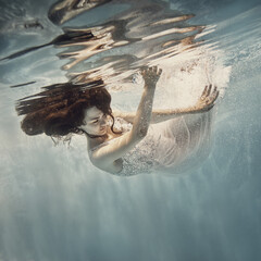 Wall Mural - A girl in a white dress with dark hair swims underwater as if flying