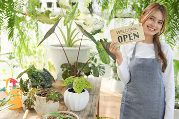 A woman selling a tree holds a welcome open sign in the green house.