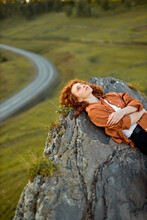 Redhead Curly Woman Is Lying On Large Rock At High Altitude. View Of Nature And Greenery In The Background. Adorable Lady Enjoys Spending Time In Nature, In Contemplation. People, Travel Concept