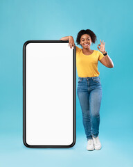 Wall Mural - Positive black lady posing near huge cellphone with empty screen, showing okay gesture, recommending website, mockup