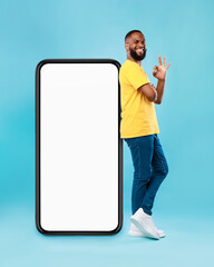 Poster - Optimistic black man showing ok gesture, leaning on big smarthone with blank screen, recommending new mobile app, mockup