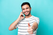 © luismolinero - Young Brazilian man isolated on blue background keeping a conversation with the mobile while doing thumbs up