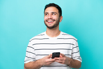 young brazilian man isolated on blue background using mobile phone and looking up