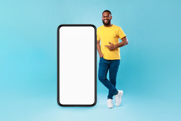 Poster - Positive Afro guy pointing at big smartphone with mockup for mobile app, recommending cool website over blue background