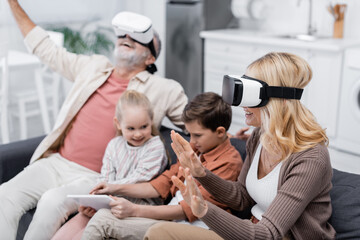 excited couple gaming in vr headsets near grandchildren using digital tablet on couch.