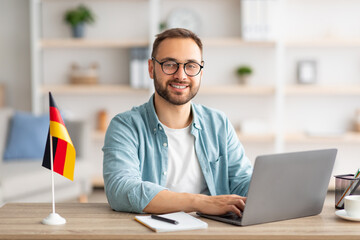 Poster - Happy young guy sitting at table with flag of Germany, using laptop pc, studying foreign language online at home