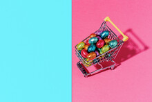 A Shopping Cart With Colourful Chocolate Eggs On A Pink And Blue Background.