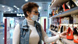© emaria - Elderly woman in medical mask and eye glasses choosing shoes in a shoe store. Shopping mall, safety during coronavirus pandemic.