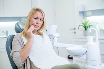 Middle shot of a distressed Caucasian woman having a toothache, sitting in a dental chair, and touching her cheek.