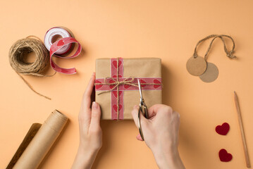 Wall Mural - First person top view photo of valentine's day decor female hands cutting twine on craft paper giftbox with scissors roll of kraft paper spool of ribbon pencil tags hearts on isolated beige background