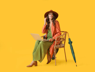 Wall Mural - Young fashionable woman sitting in chair with laptop on yellow background