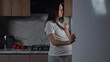 © lenblr - Pregnant brunette woman with long hair wearing pajamas eats greenery in kitchen at late night stroking belly illuminated with fridge light.