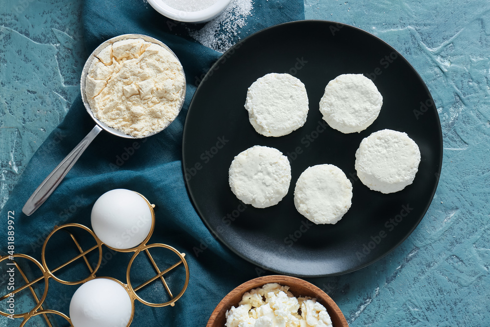 Plate with uncooked cottage cheese pancakes and ingredients on blue background