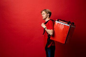 Wall Mural - Young delivery employee man in red tshirt uniform with a thermal food bag backpack works as a courier in a food delivery service on a white background.