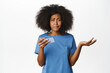 © Cookie Studio - Confused african american woman shrugging, holding smartphone, staring puzzled at camera, standing in blue t-shirt over white background