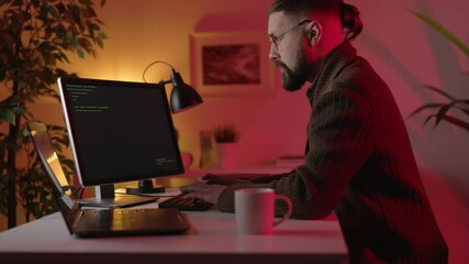 Caucasian man in eyeglasses and casual wear sitting at workplace and writing code on two modern computers. Concept of programming, technology and IT sphere.