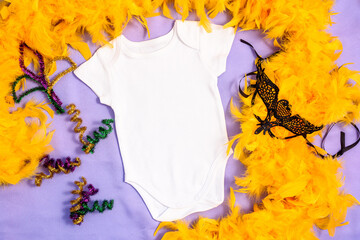 Mockup white baby bodysuit decorated yellow feather boa and carnival mask. First Mardy Gras baby apparel flatlay on fabric lilac background, flat lay, top view, copy space.