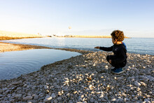 Squatting Boy Throwing A Stone Into The Water On The Seashore