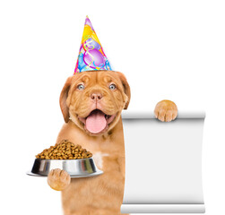 Poster - Happy Mastiff puppy wearing a party cap holds  bowl of dry dog food and shows empty list. isolated on white background