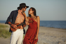Happy Couple In Love Sensual Portrait. Caucasian Man Playing Ukulele Serenading And Girl In Red Dress Dancing On The Beach At Sunset. A Bearded Handsome Man And Stunning Woman Having Romantic Tender