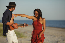Happy Couple In Love Sensual Portrait. Caucasian Man Playing Ukulele Serenading And Girl In Red Dress Dancing On The Beach At Sunset. A Bearded Handsome Man And Stunning Woman Having Romantic Tender