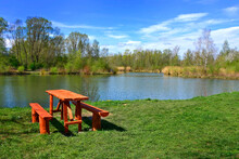 Wooden Table And Benches On A Lake Coast. Green Grass And Calm Water Surface. Relaxation Area.