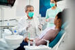 Mature dentist with face mask talks to black female patient at dentist's office.