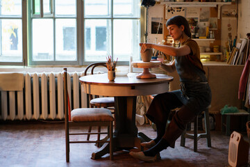 Poster - Creative ceramist molding clay vase sitting at table wearing dirty apron during master class in studio. Young businesswoman, pottery studio owner making handmade ceramics for holiday sale in workshop