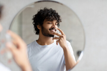 Satisfied indian man smelling perfume, doing morning beauty routine, enjoying pleasant odor, standing in bathroom