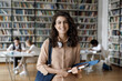 © fizkes - Happy Hispanic gen Z student girl with headphones visiting public library for work on study research project, holding learning papers, notebook, looking at camera, smiling. Head shot portrait