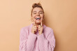 Positive young woman with combed hair points at her broad toothy smile feels very happy has white perfect teeth wears knitted sweater isolated over beige background. People and emotions concept