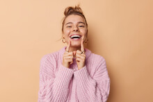 Positive Young Woman With Combed Hair Points At Her Broad Toothy Smile Feels Very Happy Has White Perfect Teeth Wears Knitted Sweater Isolated Over Beige Background. People And Emotions Concept