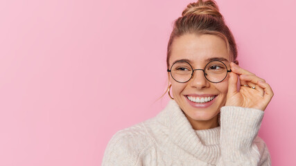 Wall Mural - Portrait of cheerful young pretty woman keeps hand on rim of spectacles smiles broadly hears good news focused away wears white sweater isolated over pink background blak copy space for promotion