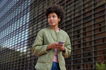 Wall Mural - Young curly haired woman looks away thoughtfully dressed in jacket holds mobile phone connected to wireless internet walks outdoors strols in town uses navigation checks route. Technology concept