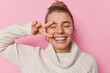 Portrait of good looking young woman smiles broadly shows white teeth makes peace gesture over eye has hair combed in bun wears warm winter sweater isolated over pink background. Stay positive