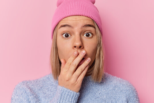 Shocked embarrassed young woman has short hair covers mouth with hand hears something unexpected and breathtaking wears hat and casual soft jumper poses against pink background. Omg concept.