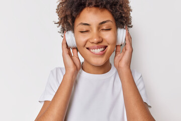 Wall Mural - Happy female meloman enjoys sound in new headphones keeps eyes closed bites lips listens favorite music dressed casually isolated over white background. People hobby and modern technologies.
