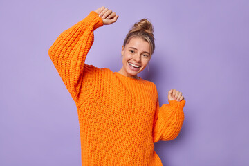 Wall Mural - Happy carefree European woman smiles gladfully moves with rhythm of music shakes arms has fun wears knitted sweater isolated over purple background catches every bit of song has upbeat mood.
