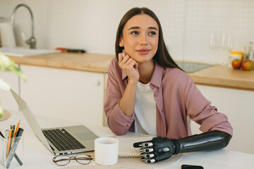 Beautiful pretty female cyborg with artificial 3d-printed arm prosthesis making wish list holding pen in hand, looking up with thoughtful pensive face expression sitting in kitchen next to laptop