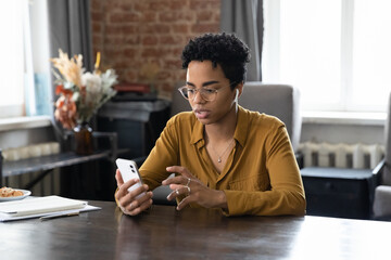 Serious focused Black millennial business woman in glasses using online app on cellphone at home office workplace table, looking at phone screen, reading text message, browsing internet on smartphone