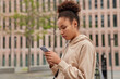 © WHstudio Leushin N - Sideways shot of curly sporty woman dressed in hoodie uses mobile phone chats online checks message content goes to gym poses against urban background has serious expression. Sport and technology