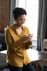 Happy African millennial professional girl in glasses reading text message on smartphone, smiling, feeling joy, chatting, typing, using online service, virtual app on internet in home office