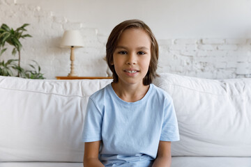 Positive pre teen gen Z boy looking and speaking at camera on video call from home. Schoolchild kid sitting on white couch in living room head shot portrait. Screen view