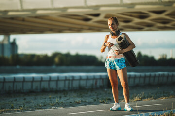 Wall Mural - Woman With Rolled Up Exercise Mat And Water Bottle Resting After Training Near The River