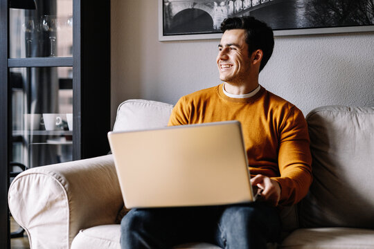 happy hispanic man using laptop in sofa at home, mustard color